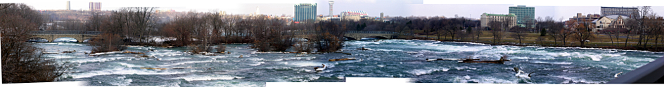 [Eight photos stitched together to give a panoramic view of the rapids and the islands with leafless trees in the rapids. There are some buildings along the shoreline on the right and in the far distance in the center on the other side of the Falls.]